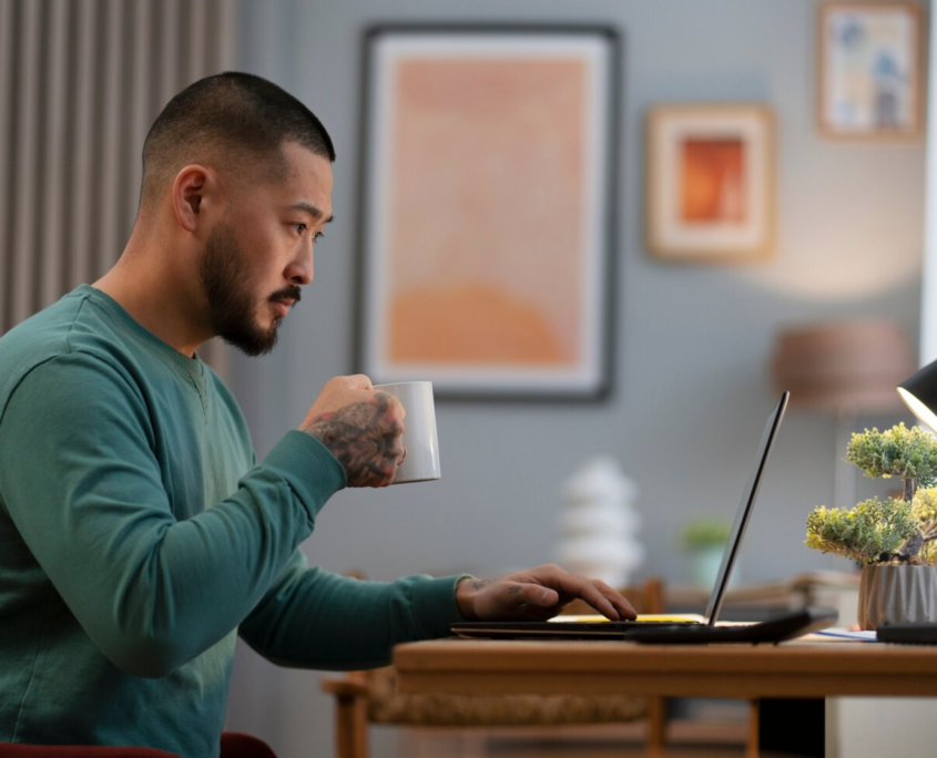 An Asian man working at a desk from home holding a mug of coffee