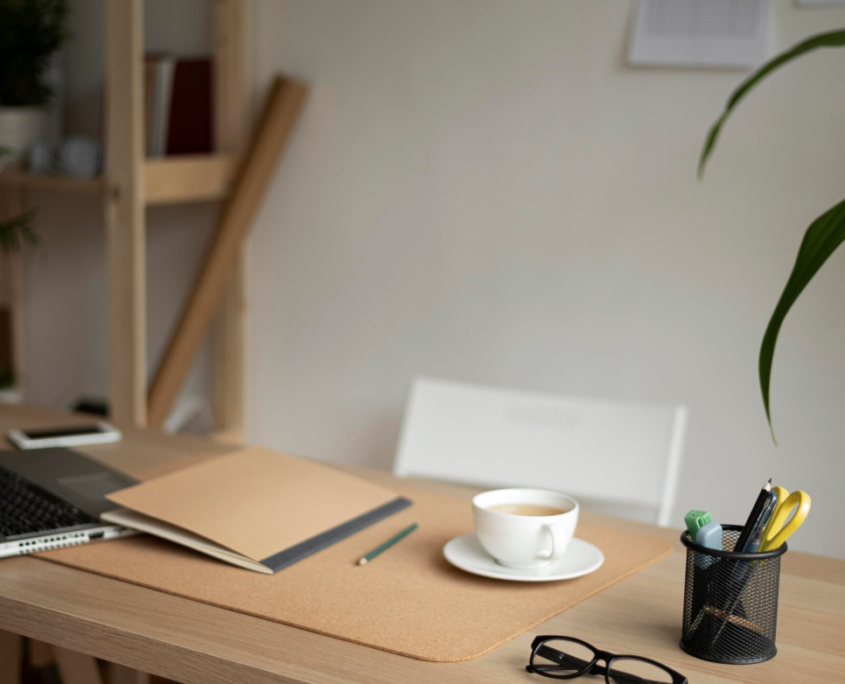 A tidy desk with no clutter, a laptop, notebook and coffee cup with shelving in the background